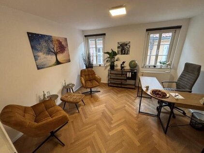 „Beratungsraum mit Fischgrätparkett, weißen Wänden und großen Fenstern. Einrichtung: Holzschreibtisch, grauer Bürostuhl, zwei braune Lounge-Sessel mit Fußhockern, dekorierte Kommode, Wandbilder und Pflanzen. Atmosphäre: ruhig und professionell.