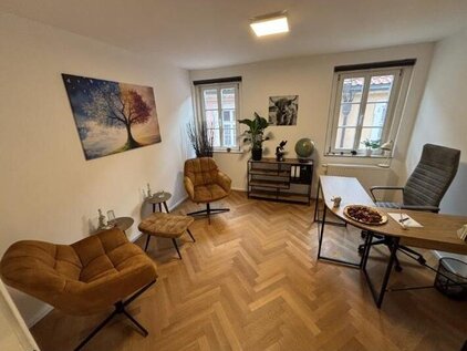 „Beratungsraum mit Fischgrätparkett, weißen Wänden und großen Fenstern. Einrichtung: Holzschreibtisch, grauer Bürostuhl, zwei braune Lounge-Sessel mit Fußhockern, dekorierte Kommode, Wandbilder und Pflanzen. Atmosphäre: ruhig und professionell.