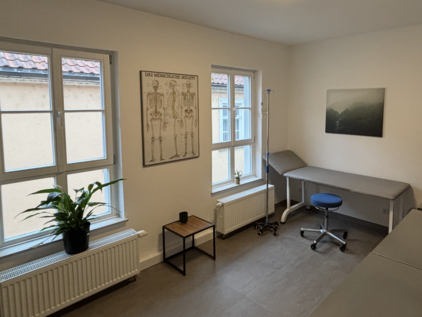 Sprechzimmer mit Untersuchungsliege, blauem Rollhocker, Infusionsständer und Wandposter ‚Das menschliche Skelett‘; helle Fenster, Holzfußboden und dezente Dekoration sorgen für eine ruhige, medizinische Atmosphäre.