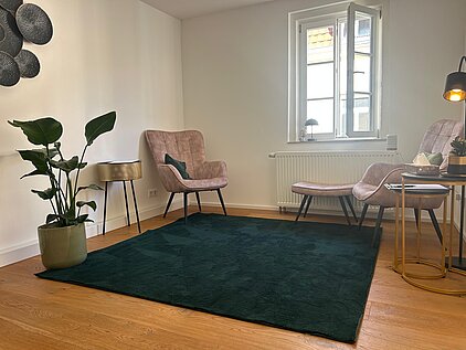Gemütliches Sprechzimmer mit zwei rosa Sesseln, schwarzem Teppich, Beistelltischen und Wanddekoration in heller, entspannter Atmosphäre.